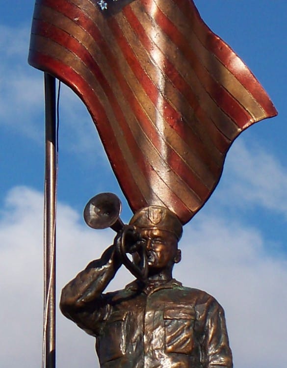 Bronze sculpture of soldier playing taps with half-mast American flag.