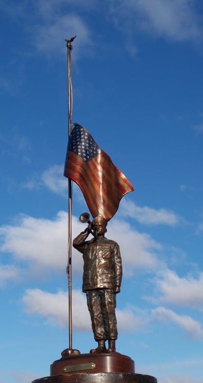 Bronze sculpture of soldier playing taps with half-mast American flag.