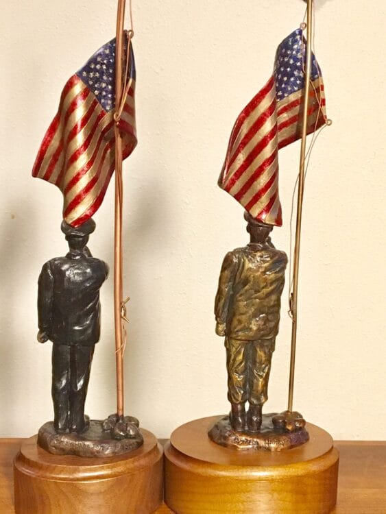 Back view of two sculptures of an American soldiers playing taps beside an American flag.