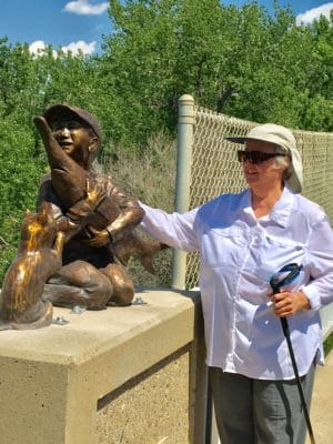 photo of the artist Pamela Harr posing with a large bronze sculpture of a little boy, his cat, and a paddlefish.