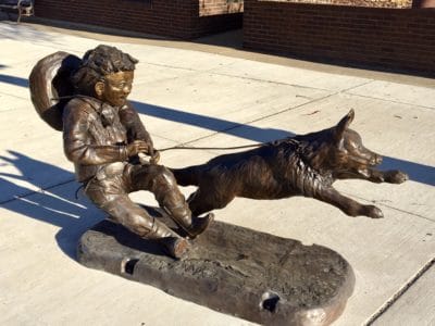 Bronze sculpture of a dog on a leash pulling a young boy who is trying to stop him.